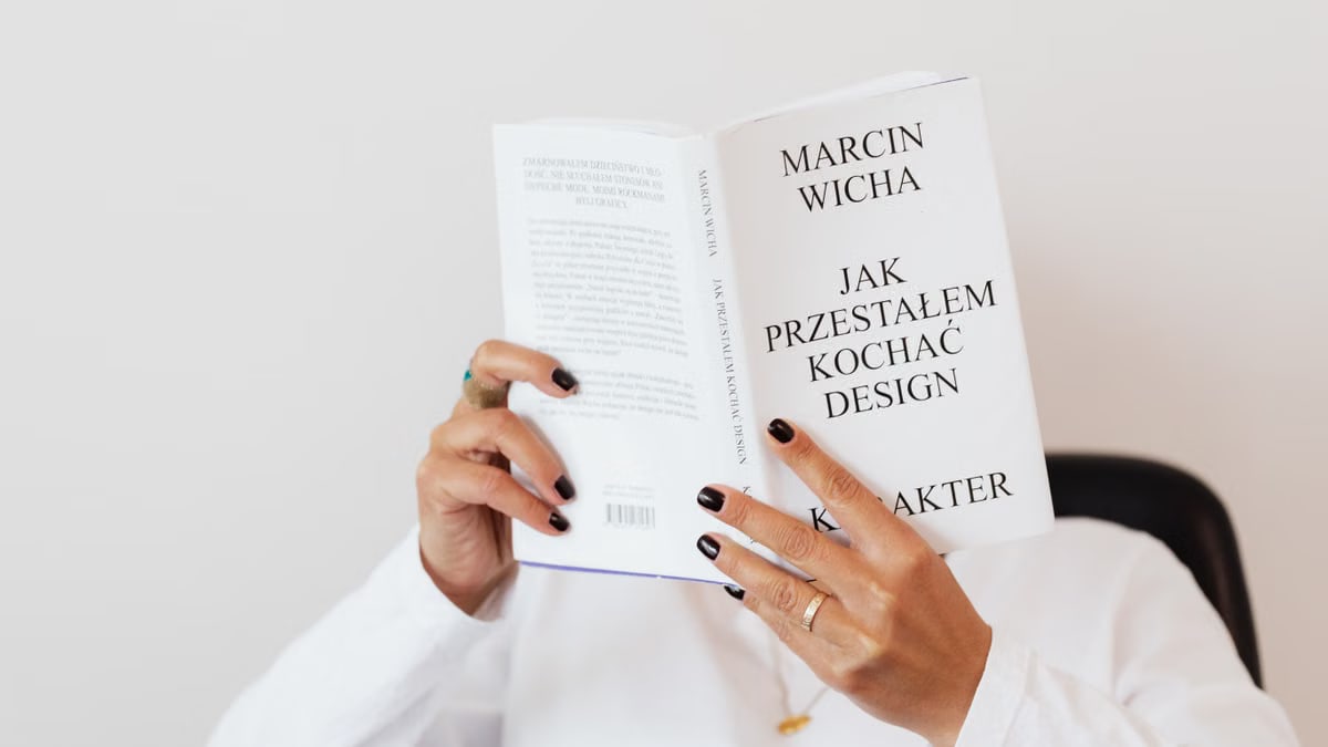 Anonymous female in white shirt with gold chain on neck reading interesting book covering face while sitting on black chair